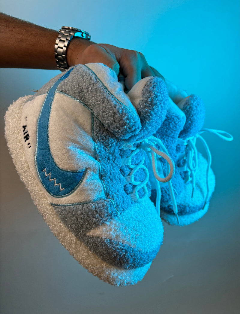 Close-up of a hand holding a pair of blue and white Cozify fuzzy slippers with soft sherpa texture and sneaker-inspired design under cool lighting.