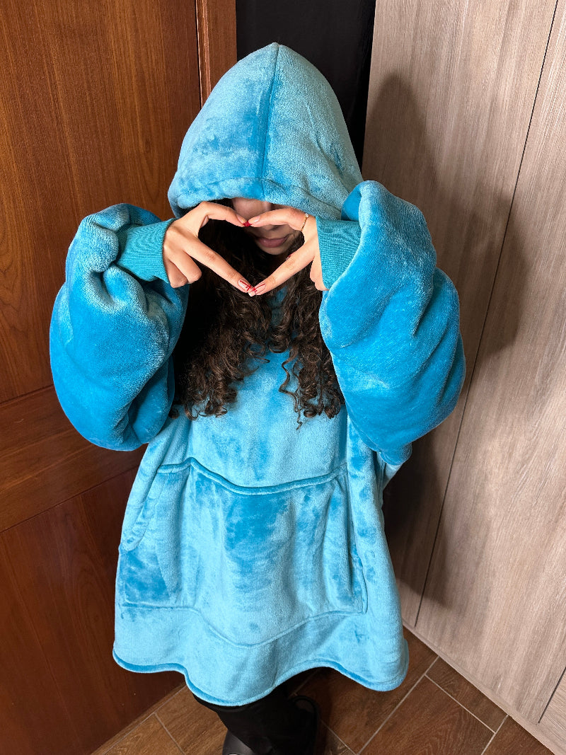 A woman wearing a blue Cozify oversized wearable blanket hoodie stands with the hood up, forming a heart shape with her hands in front of her face.