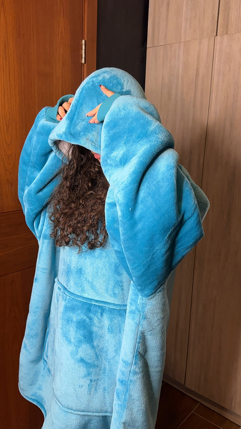 A person wearing a blue Cozify oversized wearable blanket hoodie pulls the hood over their head, showing the plush texture and loose fit of the cozy fabric.