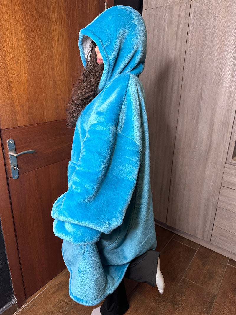 A woman wearing a bright blue Cozify oversized wearable blanket hoodie stands in a side view, showing the hood and plush sherpa texture against a wooden door background.