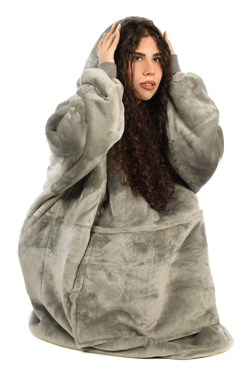 A woman sitting on the floor wearing a gray Cozify plush wearable blanket hoodie and matching gray slippers, facing forward and showing the soft, oversized design and cozy texture.