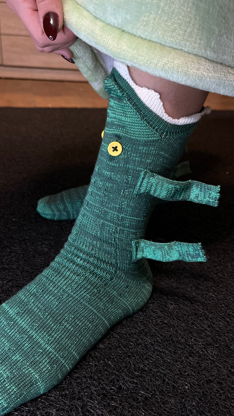 A close-up side view of a person wearing green knit Cozify socks featuring yellow button accents and ribbed textured fabric, shown against a dark carpet background.