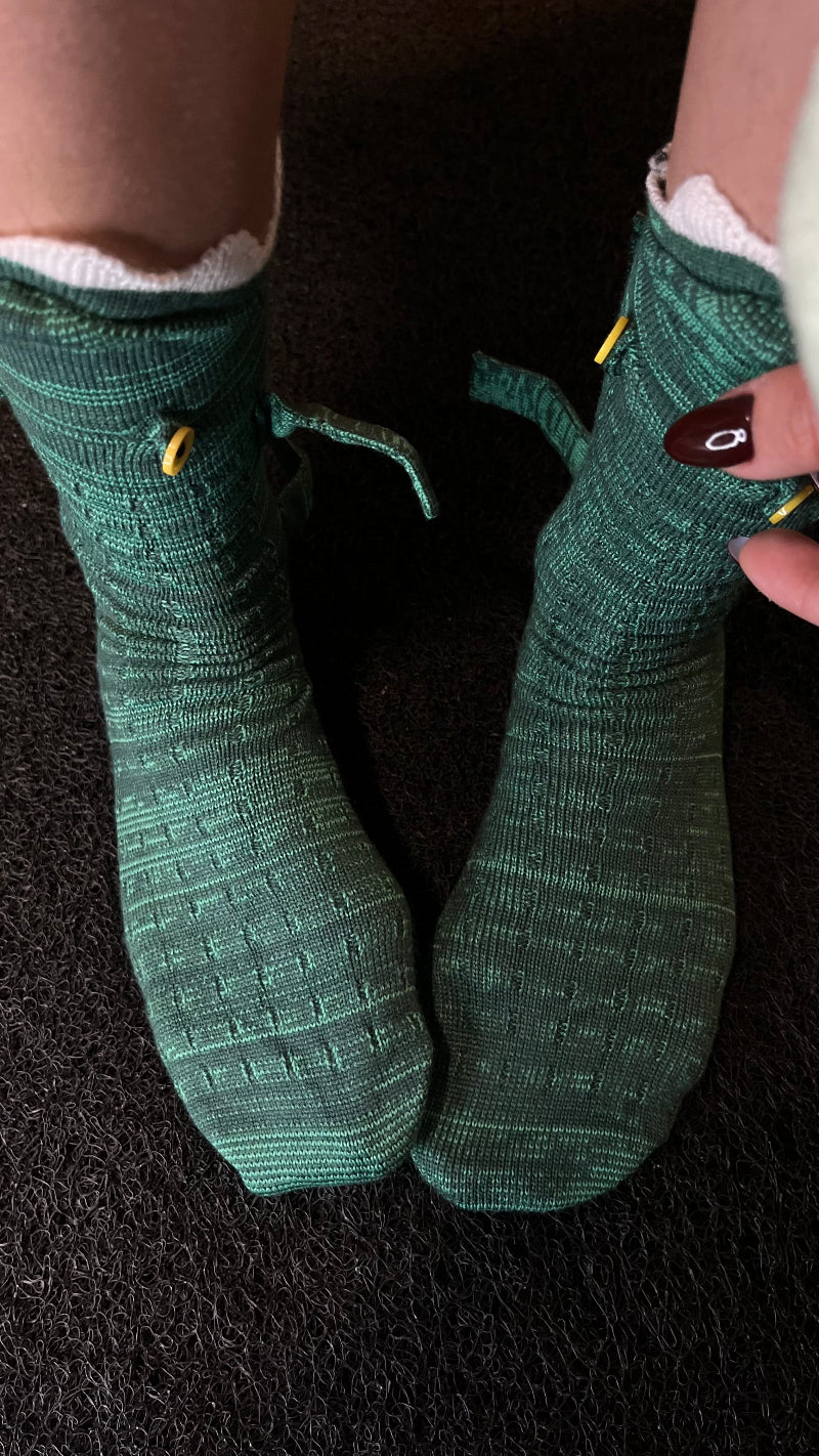 A close-up view of a person wearing green textured knit Cozify socks with small yellow details, shown against a dark carpet background.