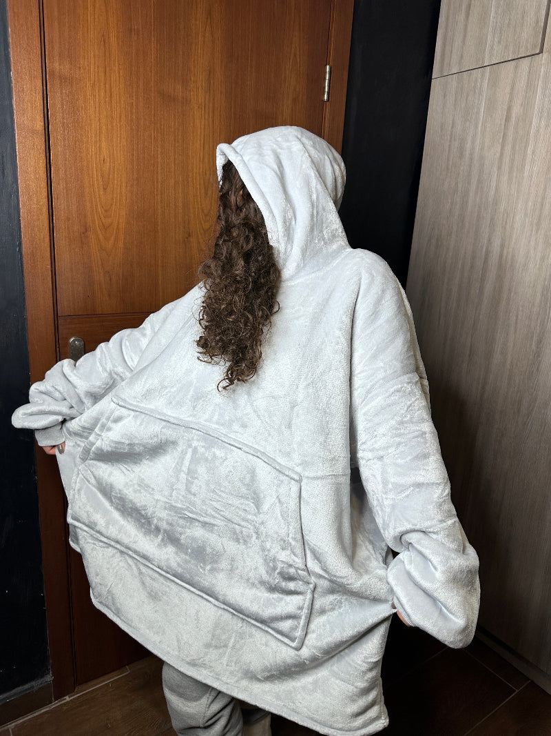A woman wearing a light gray Cozify plush wearable blanket hoodie gently holds the hood over her face, showing the soft and cozy texture of the oversized design.