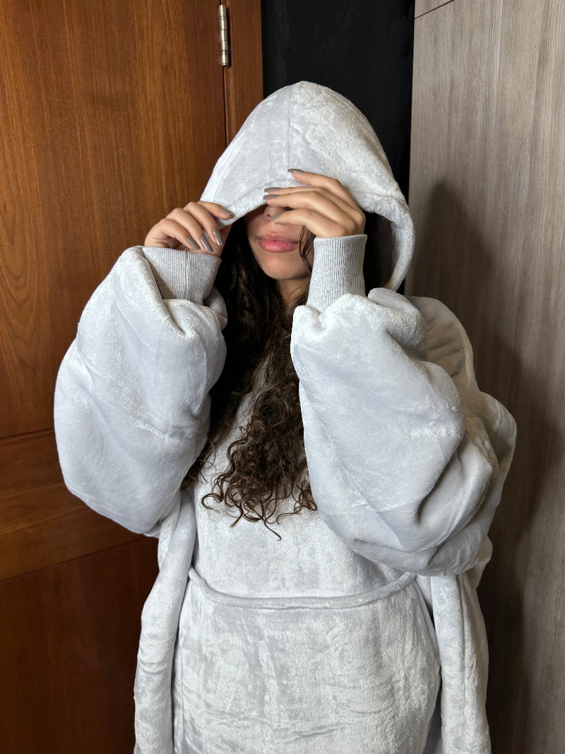A woman wearing a light gray Cozify plush wearable blanket hoodie gently holds the hood over her face, showing the soft and cozy texture of the oversized design.