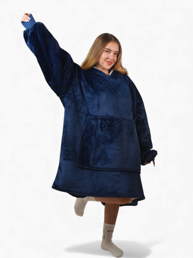 Smiling woman in a full-body view wearing a navy blue oversized Cozify sherpa-lined hoodie blanket with a front pocket, raising one arm playfully.
