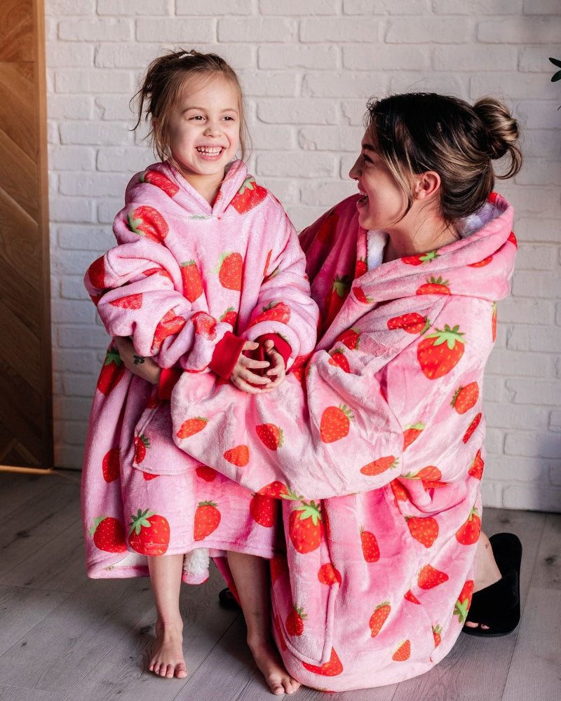 A mother and daughter smiling and wearing matching Cozify wearable blanket hoodies in pink with an all-over red strawberry print. The oversized sherpa-lined hoodies look cozy and soft, with the mother kneeling and the daughter standing barefoot indoors against a white brick wall.