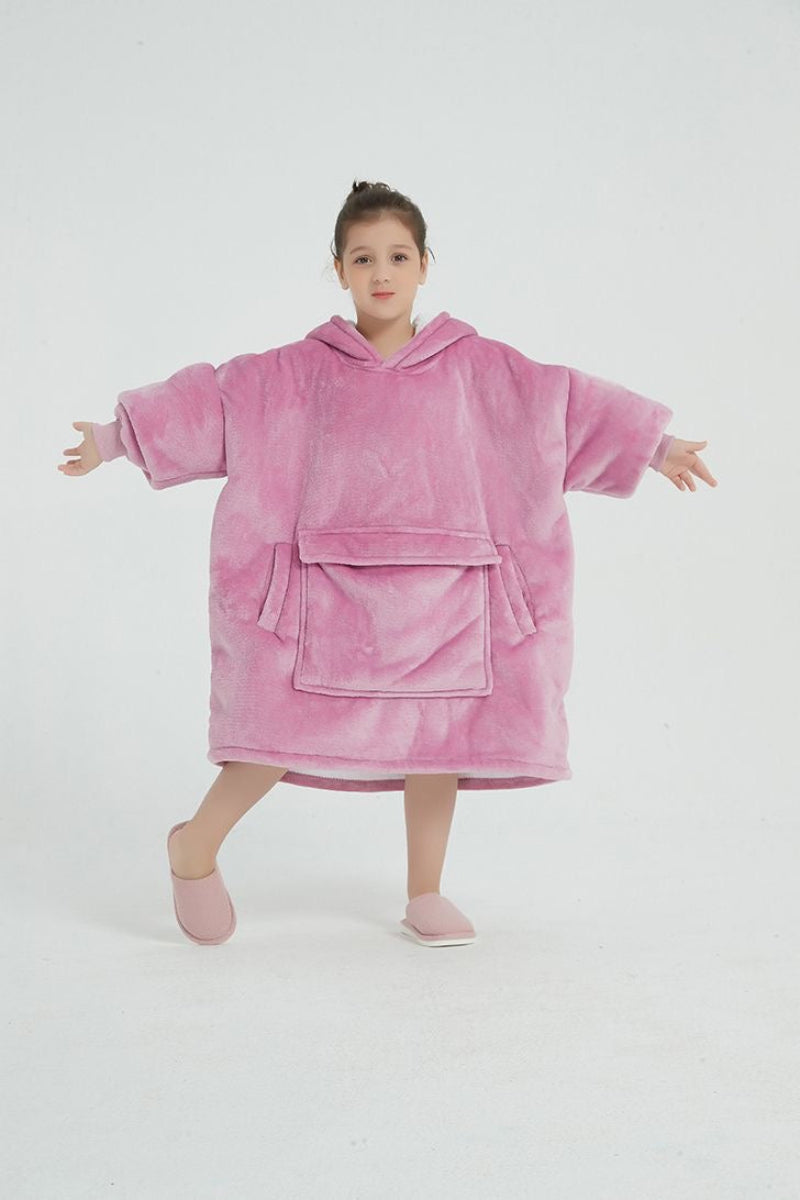 A young girl poses in a bright pink oversized hoodie blanket with a front kangaroo pocket and hood. The fabric appears plush and cozy, designed for comfort and warmth. She is wearing matching pink slippers and standing against a plain white background, with one leg lifted and arms stretched outward playfully.