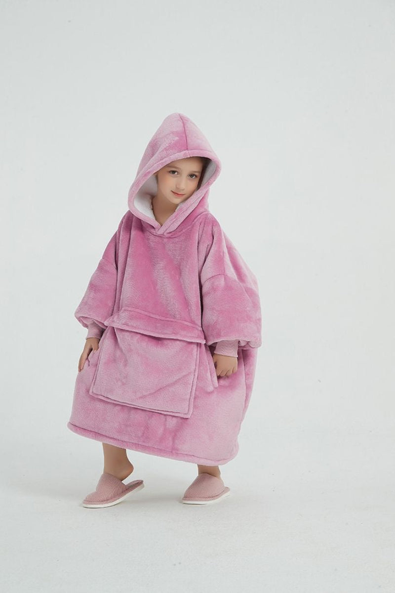 A young girl wearing a pink oversized plush hoodie with the hood up. The hoodie features a large front pocket and long sleeves. She is smiling slightly and wearing pink slippers, standing against a plain white background.