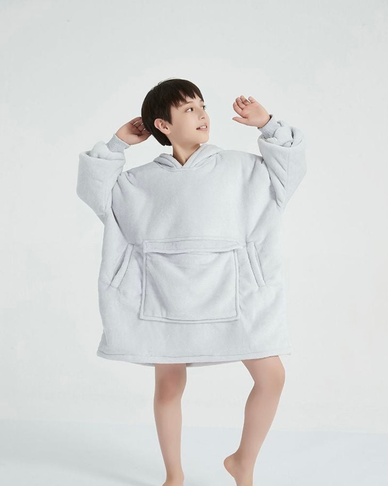 A child wearing an oversized, plain white hoodie with a large front pocket and loose sleeves. The child is barefoot, standing in a playful pose, looking upward with arms raised, emphasizing the comfort and roominess of the garment.