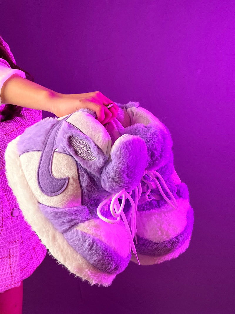 Close-up of a pair of Cozify oversized plush sneaker-style slippers in cream and purple tones, held in hand under a pink-purple spotlight. Features faux fur texture and lace-up design.