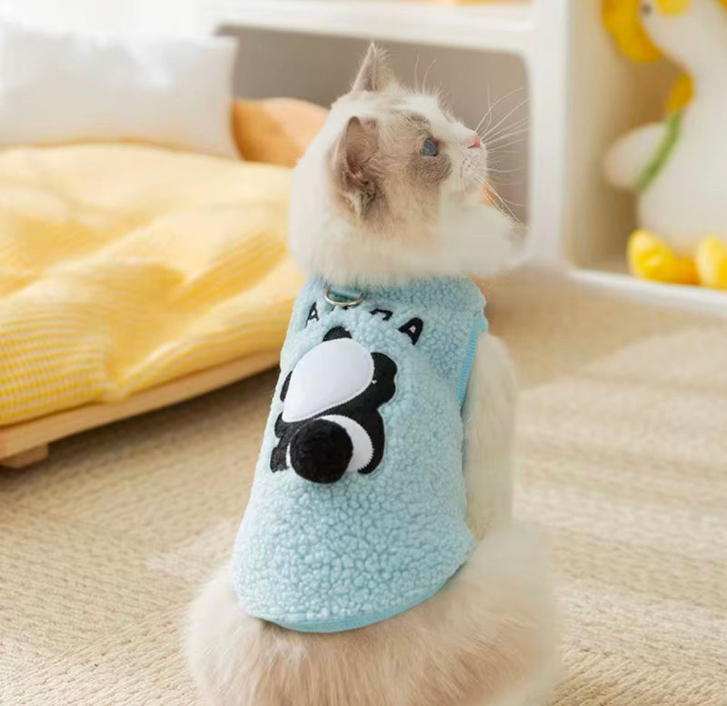 White cat wearing a light blue sherpa fleece vest with a black and white panda design on the back, sitting on a beige carpet and facing sideways. The cozy indoor setting includes a yellow pet bed and soft furnishings.