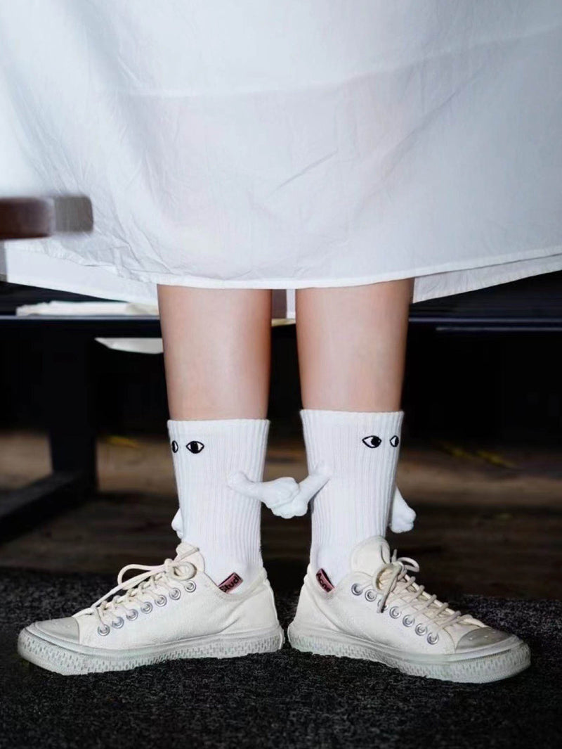 A person wearing white Cozify socks with eye designs and small 3D arms paired with cream sneakers, shown from behind under a white dress.