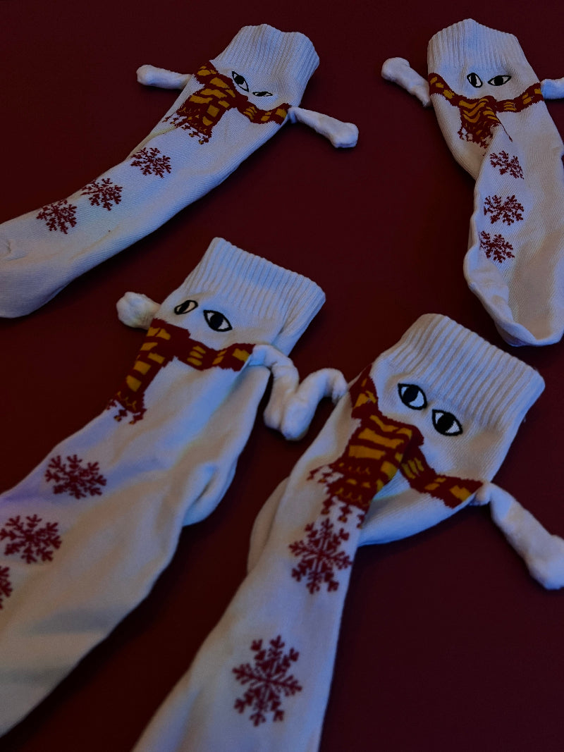A flat lay of four pairs of white Cozify socks featuring red snowflake patterns and yellow-red scarf designs on a dark red background.
