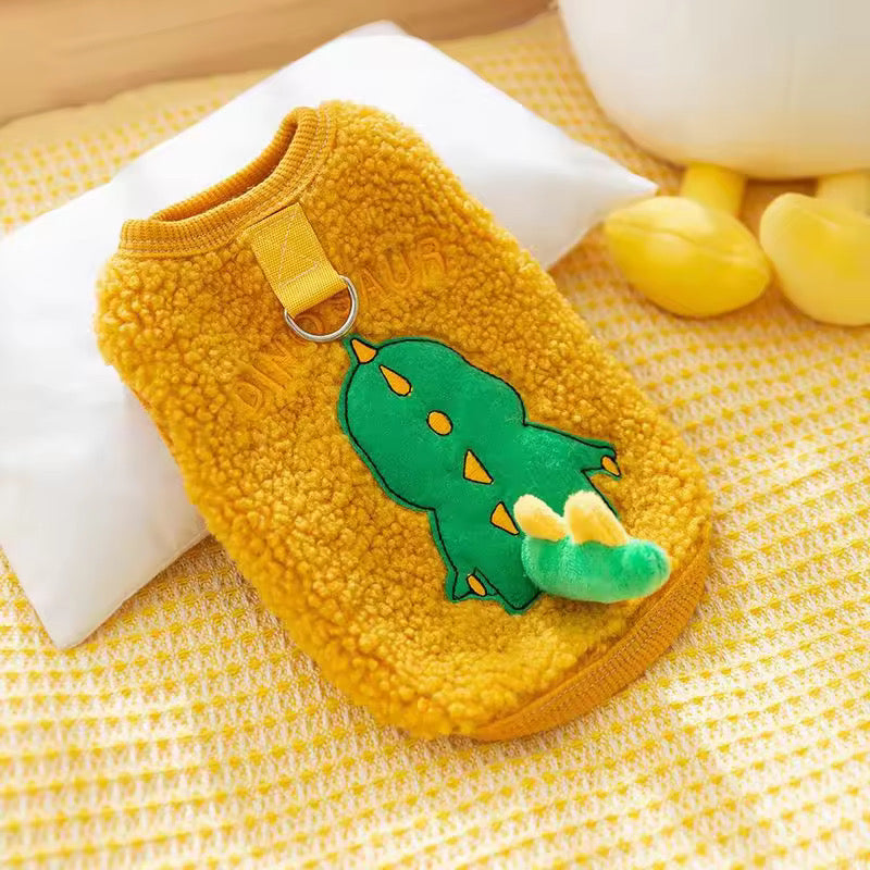 Flat lay of a mustard yellow sherpa fleece pet vest by Cozify, featuring a playful green dinosaur patch with plush tail detail and the word "DINOSAUR" embroidered above, with a yellow leash ring attachment at the neck, shown on a yellow textured background.