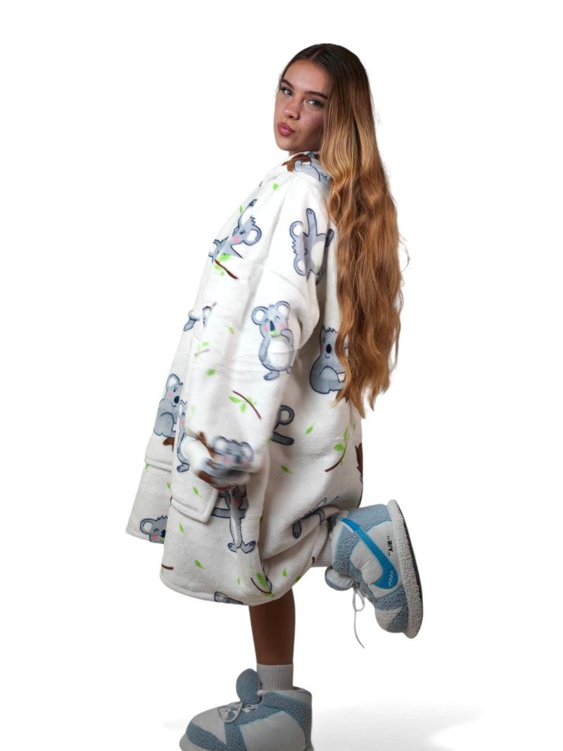 Woman wearing an off-white oversized wearable blanket hoodie with a cute koala print and a large front pocket, posing playfully on one leg against a white background.